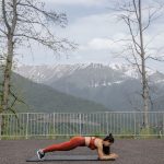 A woman doing a plank exercise on a mat outdoors with a scenic mountain view in the background.